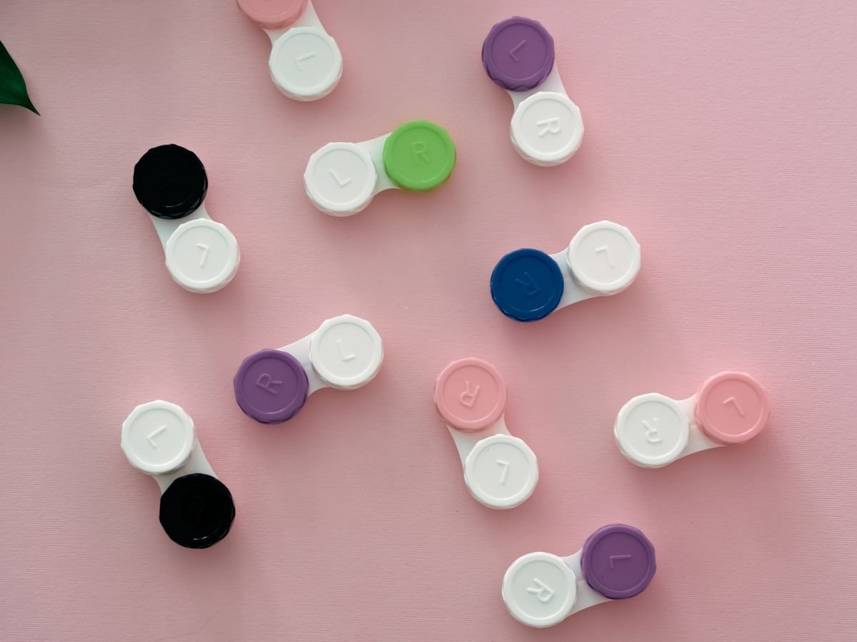 Mehrere Kontaktlinsen-Behälter in Weiß, Schwarz, Violett, Blau und Rosa auf rosa Untergrund