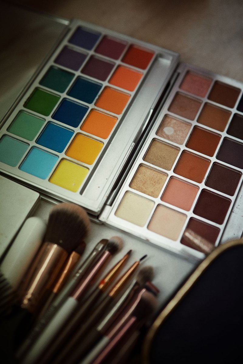 Zwei geöffnete Lidschattenpaletten in Regenbogenfarben und Erdtönen, darunter mehrere Make-up-Pinsel